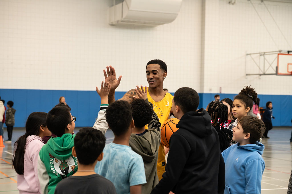 A basketball player in a yellow jersey smiles and high-fives a group of children in a gymnasium.