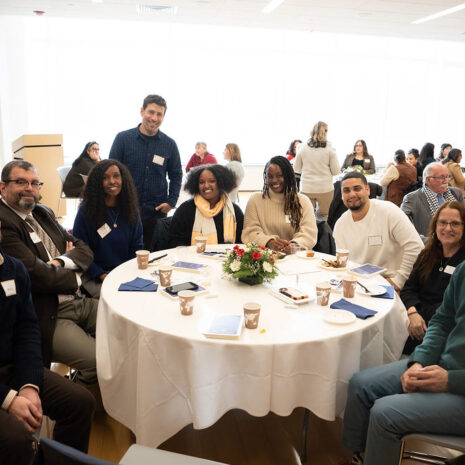 A group of nine people sit and stand around a round table set with papers, cups, and a centerpiece at a conference or meeting in a bright room.
