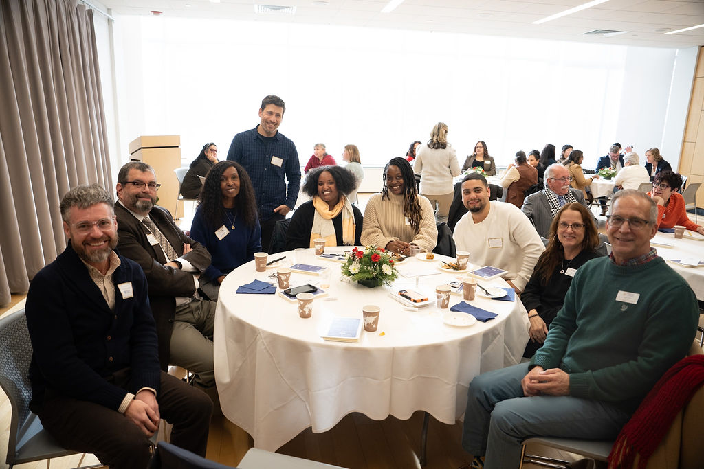 A group of nine people sit and stand around a round table set with papers, cups, and a centerpiece at a conference or meeting in a bright room.