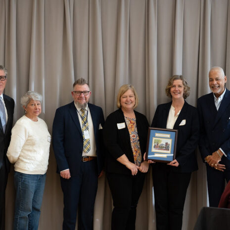 Six people stand side by side in front of beige curtains, with one person holding a framed certificate or award.