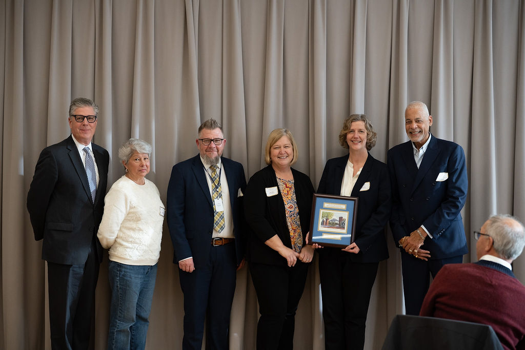 Six people stand side by side in front of beige curtains, with one person holding a framed certificate or award.
