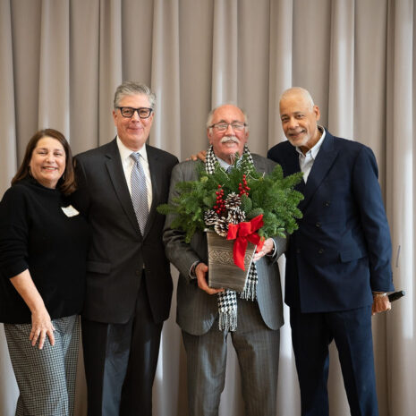Four adults pose together indoors; one man in the center holds a festive potted plant with a red bow. They are standing in front of closed beige curtains.