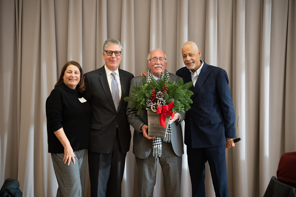 Four adults pose together indoors; one man in the center holds a festive potted plant with a red bow. They are standing in front of closed beige curtains.