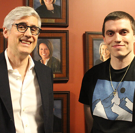Two men stand side by side in front of a wall with framed portraits. One is wearing a blazer and glasses, the other a graphic t-shirt and necklace.