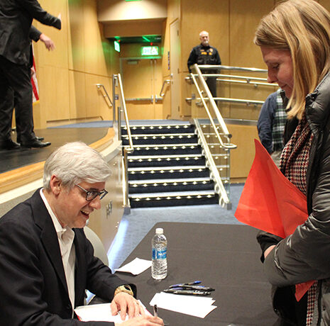 An older man sits at a table signing a book for a woman in a winter coat at an indoor event, with other people and a stage visible in the background.