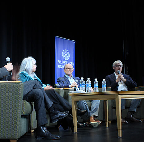 Four people sit on stage in armchairs having a panel discussion, with water bottles and papers on the table, and a university banner in the background.