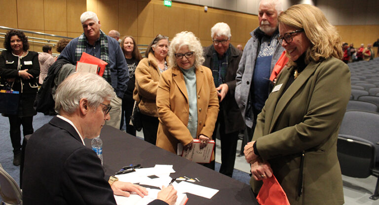A man sits at a table signing books for a group of people who are lined up and waiting in a large indoor event space.