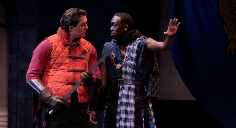 Two actors on stage, one in a red vest holding a sword and the other in a plaid costume with a raised hand, perform a dramatic scene in a theatrical production.