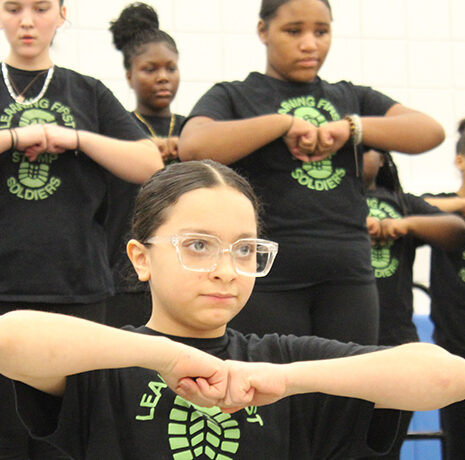 A group of young people in matching black T-shirts perform a synchronized hand gesture in a gymnasium.