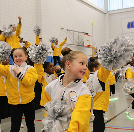 Children wearing yellow and white jackets perform a cheerleading routine with silver pom-poms in a gymnasium with basketball hoops and a WSU banner.