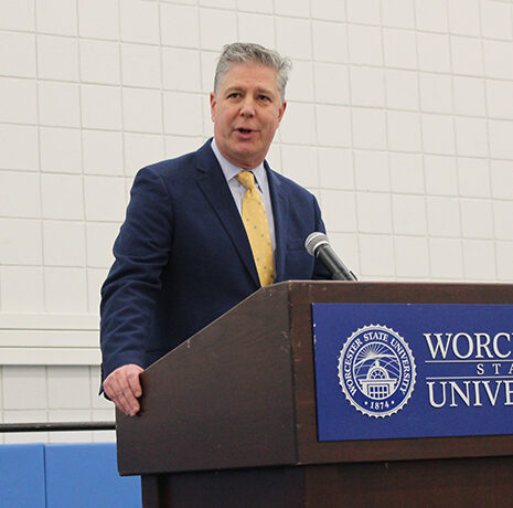A man in a suit speaks at a podium with a Worcester State University sign in an indoor setting.