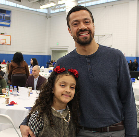A man and a young girl pose and smile in front of round tables at an indoor community event with people seated and standing in the background.