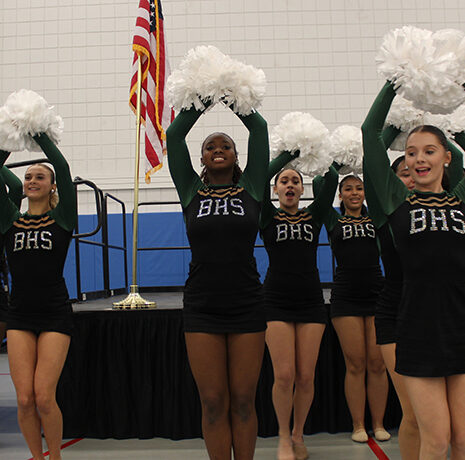 A group of cheerleaders in black and green BHS uniforms hold white pom-poms in the air while performing indoors near an American flag.