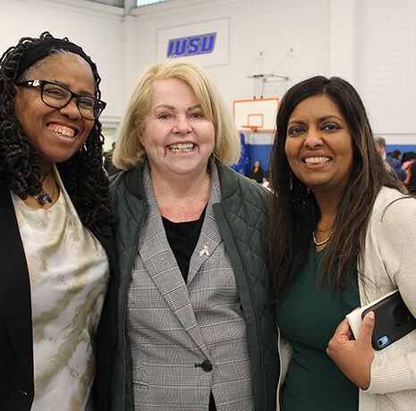 Three women stand together smiling in a gymnasium with tables, chairs, and basketball hoops visible in the background.