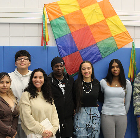 Seven people stand in a row indoors in front of a blue wall and a large, colorful, checkered kite hanging on the wall behind them.