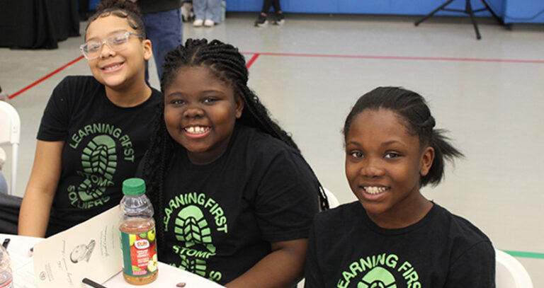 Three girls sit at a table indoors, smiling at the camera. They wear matching black “Learning First” t-shirts, and a bottle of juice and papers are on the table in front of them.