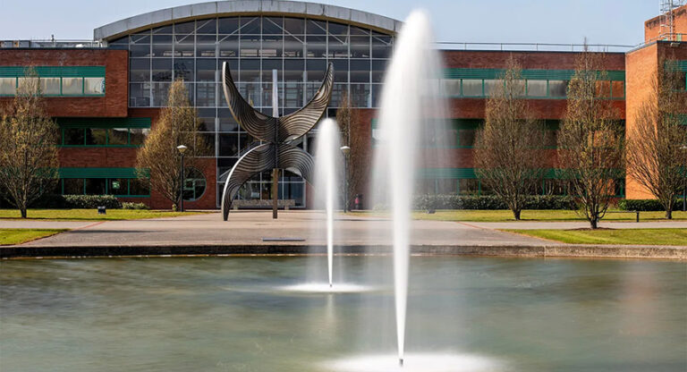 Two fountains spray water in a pond in front of a modern brick building with a large abstract metal sculpture and glass entrance.