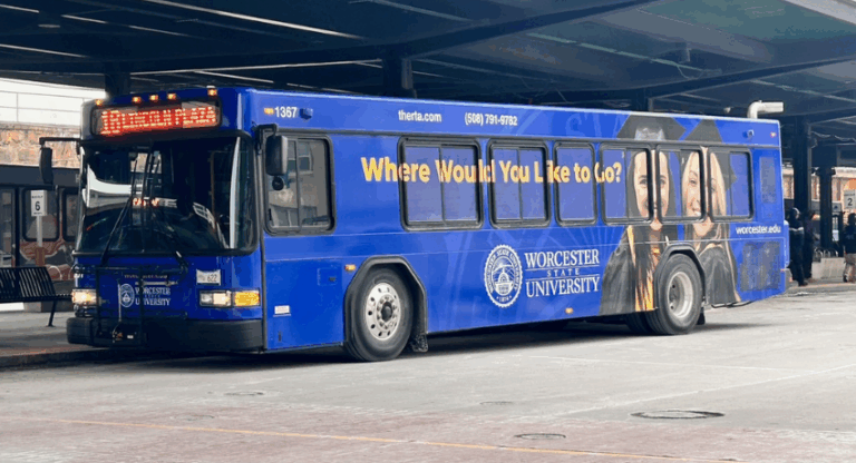 A blue Worcester State University bus is parked at a terminal, featuring an ad that reads "Where Would You Like to Go?" and university branding on its side.