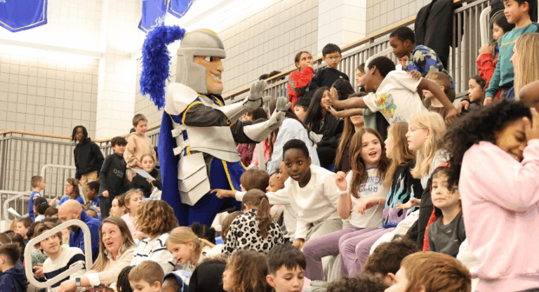 A person in a knight mascot costume interacts with children seated in gym bleachers during a school assembly.