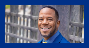 A man in a blue suit and dotted shirt smiles at the camera, standing in front of a wooden fence.