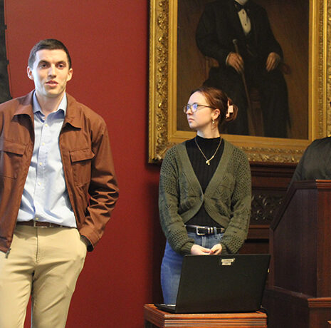 Three people stand at the front of a room giving a presentation; one speaks while holding a pointer, the other two stand behind a podium with a laptop.