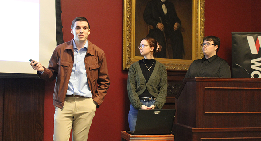 Three people stand at the front of a room giving a presentation; one speaks while holding a pointer, the other two stand behind a podium with a laptop.