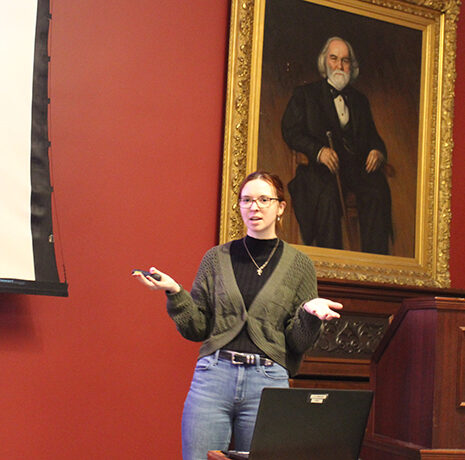 A woman stands in front of a projector screen giving a presentation in a room with red walls and a large portrait of a bearded man behind her.