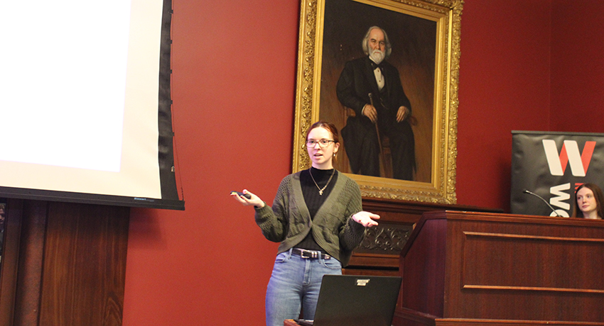 A woman stands in front of a projector screen giving a presentation in a room with red walls and a large portrait of a bearded man behind her.