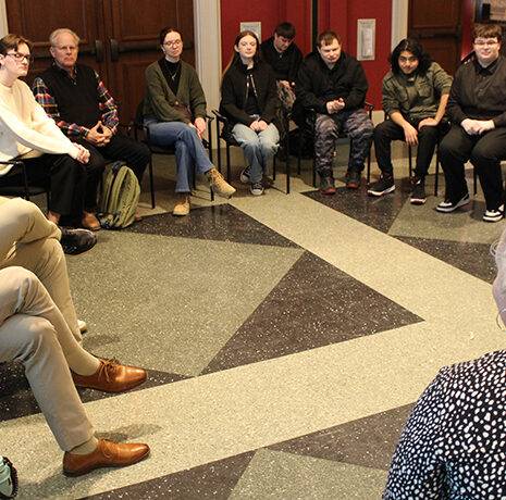 A group of people sit in a circle having a discussion in a room with red walls and double doors.