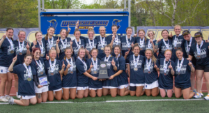 A women’s lacrosse team poses on a field, wearing medals and holding a trophy, celebrating a championship win at Worcester State University.