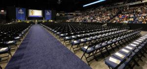 Rows of empty chairs line an indoor arena, facing a stage with blue banners and a screen, as people gather in the audience for an event.