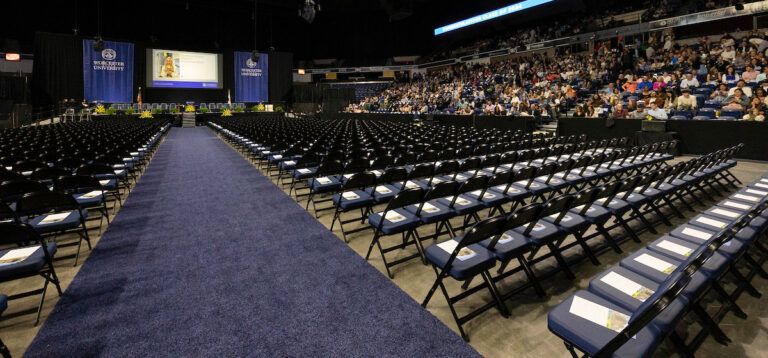 Rows of empty chairs line an indoor arena, facing a stage with blue banners and a screen, as people gather in the audience for an event.
