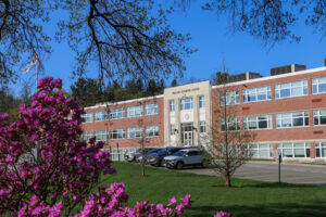 A three-story brick and white building labeled “Stamford Academic Center” sits behind a parking lot, with blooming pink flowers and trees in the foreground under a clear blue sky.
