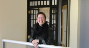 A person with short hair and a black scarf smiles while leaning on a white railing in a hallway with glass-paneled doors.