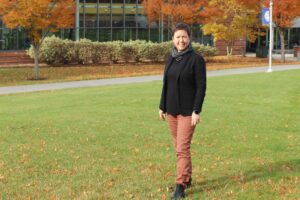 A person stands on a grassy lawn with autumn trees and a building in the background, wearing a black jacket, scarf, and rust-colored pants.