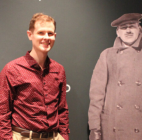 A man in a red patterned shirt stands next to a life-size black-and-white cutout of a person in a coat and cap against a dark wall with partial text.