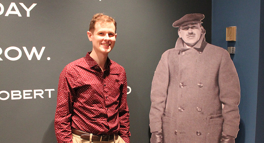 A man in a red patterned shirt stands next to a life-size black-and-white cutout of a person in a coat and cap against a dark wall with partial text.