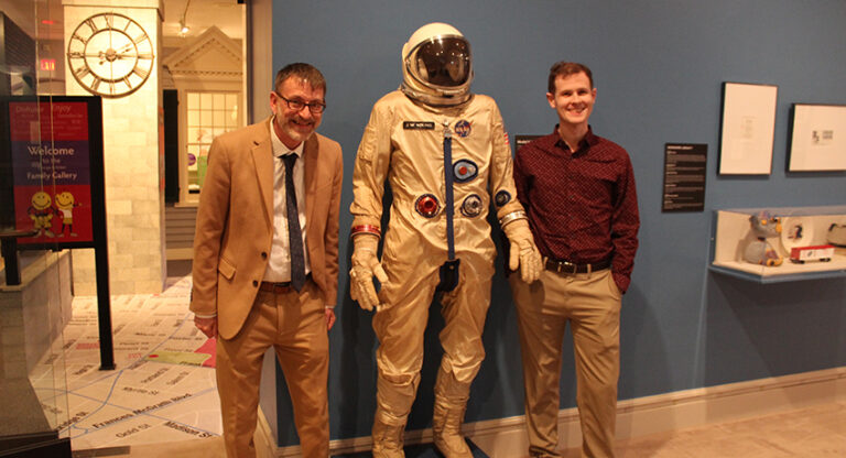Two men stand on either side of an astronaut suit display in a museum setting with blue walls and various exhibits in the background.