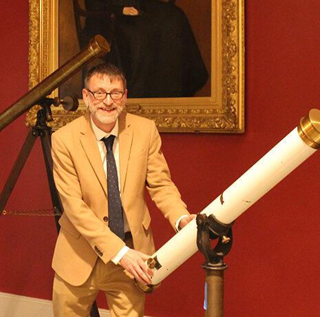 A man in a tan suit stands indoors next to a large white telescope, with a portrait, antique telescope, and American flag visible in the background.
