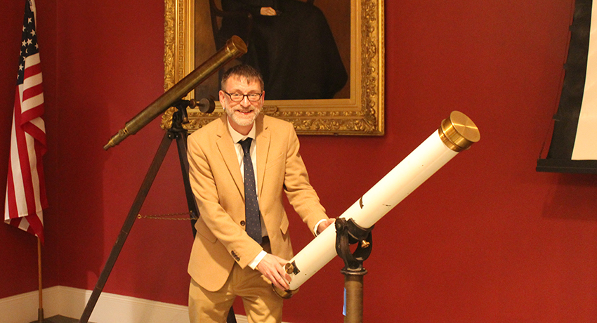 A man in a tan suit stands indoors next to a large white telescope, with a portrait, antique telescope, and American flag visible in the background.