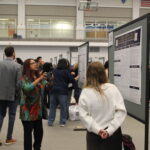 People observe and discuss research posters displayed on boards in a large indoor venue; some individuals are engaged in conversation.