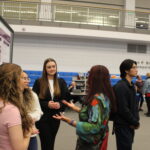 Several people stand in front of a presentation board discussing its contents at an indoor event, with others and tables visible in the background.