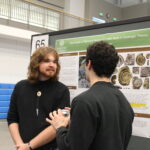 Two people stand and talk in front of a scientific poster display about fossils at an indoor event. One person holds a drink can.