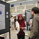A woman in a red hijab discusses her research poster with a man at an academic conference in a gymnasium setting.