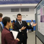 A man in a suit presents a scientific research poster to two attendees in an indoor event space with chairs and a display board.