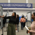 A woman presents a research poster to another woman at an academic event in a gymnasium with a "State University" banner in the background.