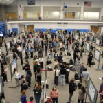 A large group of people attend a poster presentation event in a gymnasium, viewing and discussing research displays set up in rows.