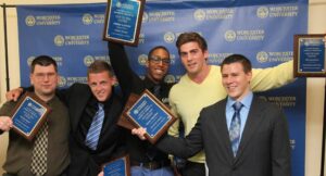 Five men pose in front of a Worcester University backdrop, each holding a plaque and smiling for the camera.