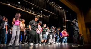 A group of people stand in a line on a stage under bright lights, following the instructions of a person pointing and speaking at the front.