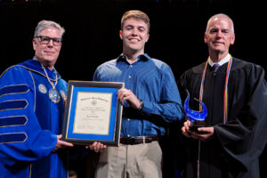 Three men stand together at an award ceremony; the man in the middle holds a framed certificate, while the man on the right holds a glass trophy.